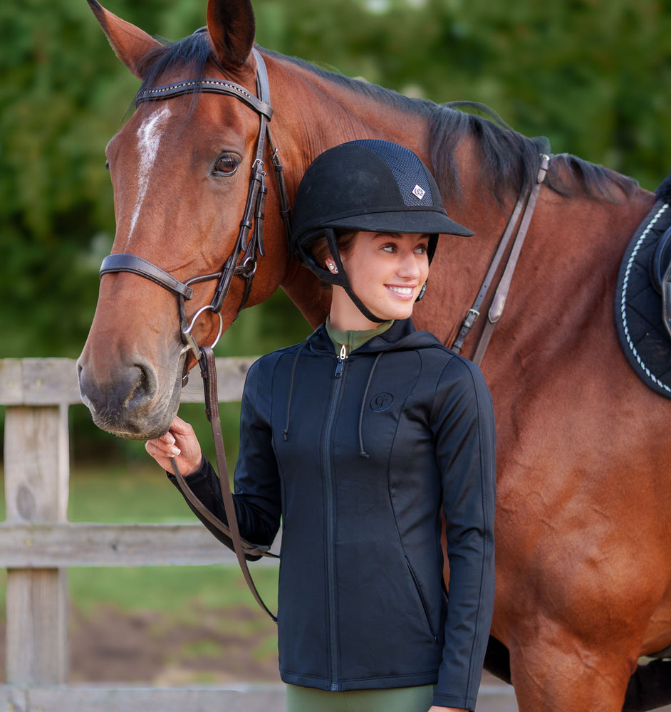 
                  
                    Woman in equestrian gear standing next to a brown horse in an outdoor setting
                  
                