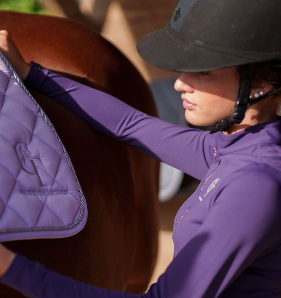 
                  
                    Close up shot of woman in purple outfit placing equestrian gear onto a brown horse
                  
                