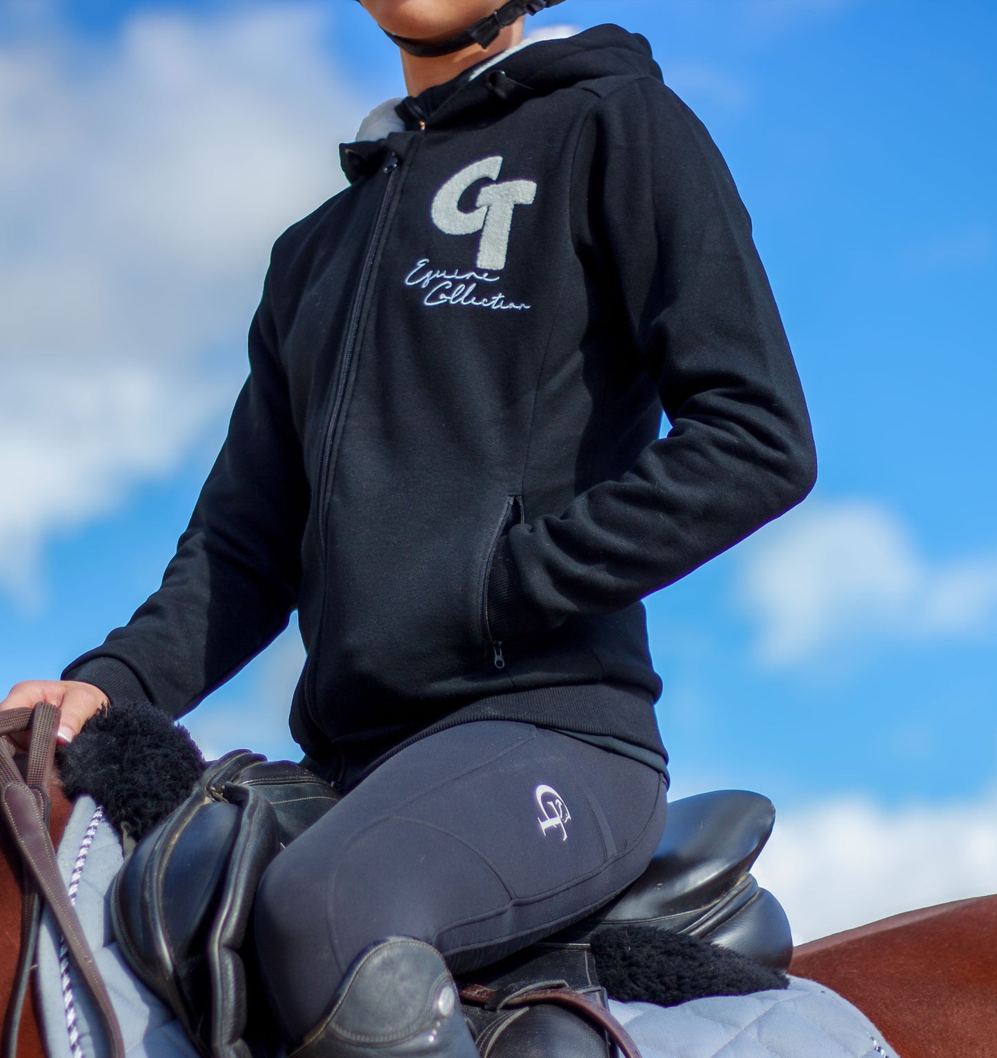
                  
                    Person riding a horse wearing a black hoodie with a logo and text, against a blue sky.
                  
                